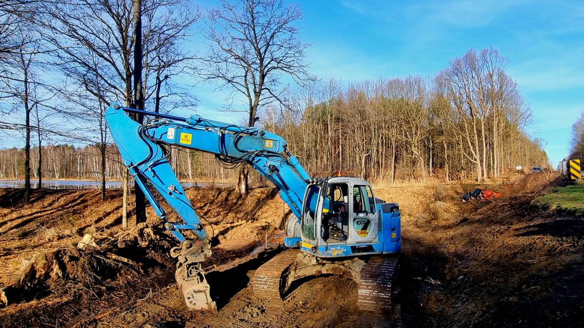 Niebieska koparka wykopuje ziemię, w tle widać jezioro i las. 