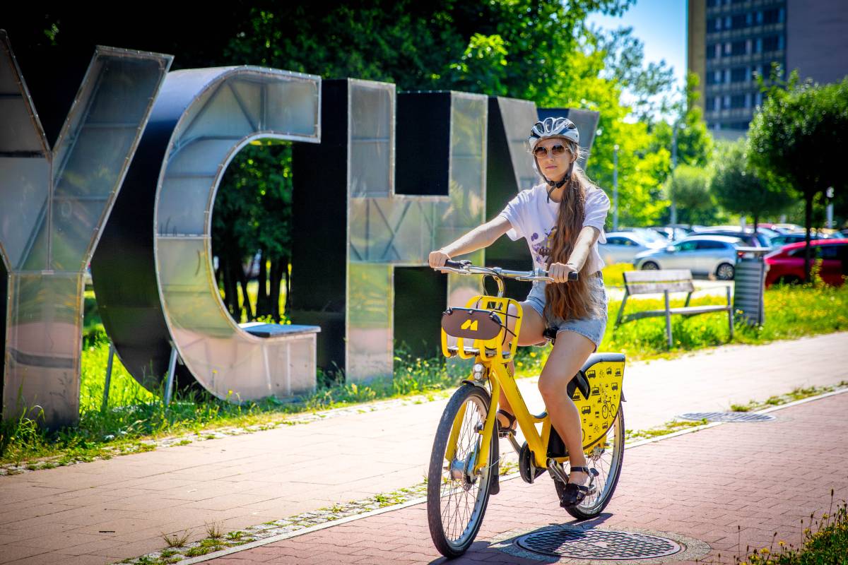Kobieta w białej koszulce z napisem "Tychy" i kaskiem na głowie, jedzie na żółtym rowerze po ścieżce rowerowej. W tle idać fragment napisu "Tychy", park, ławkę i fragment budynku.